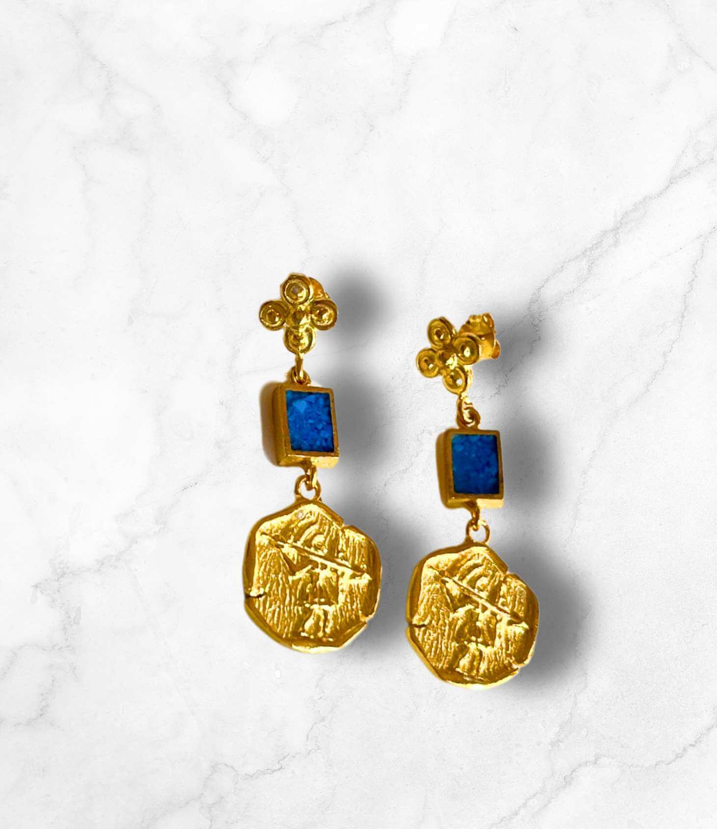Gold earrings with blue stone accents, featuring a textured disc and elegant design on a marble background.
