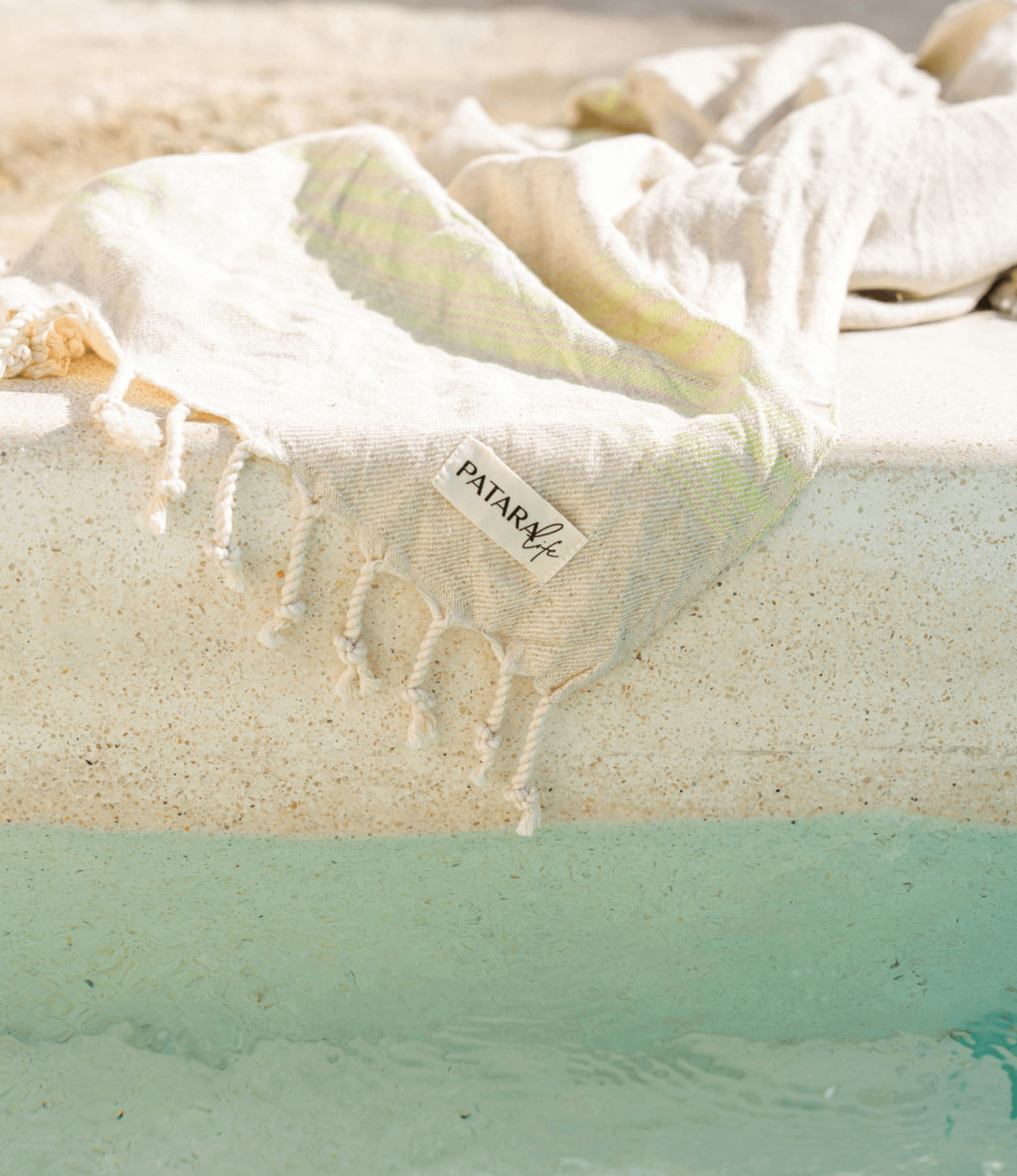 PATARA LIFE'S BEST GIFTS FOR BEACH LOVERS - a light beach towel by the poolside with a PATARA life tag.