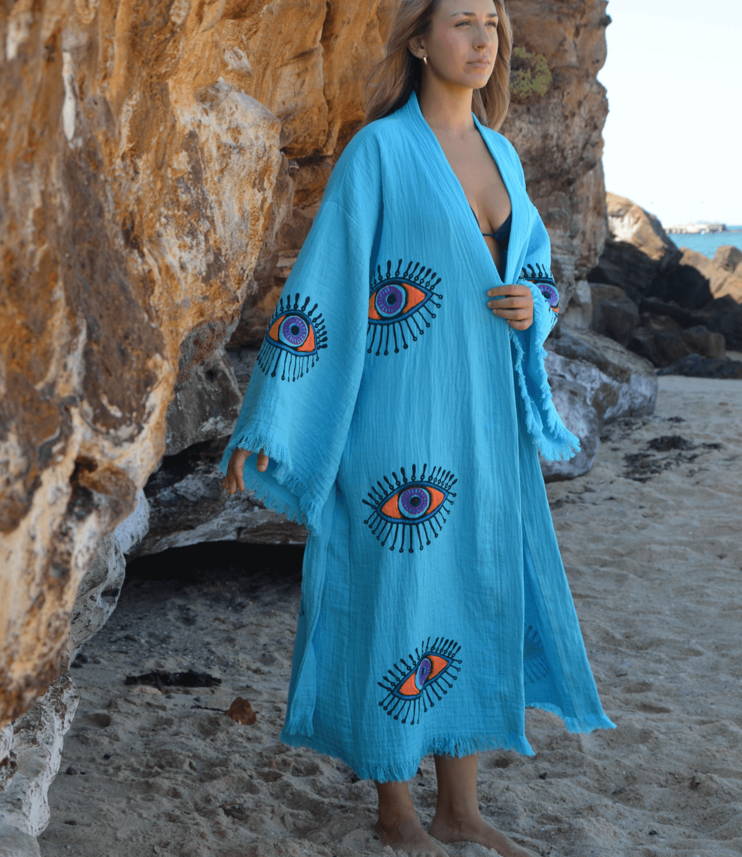 Woman wearing a turquoise Evil Eye Kaftan Robe with vibrant eye designs on a sandy beach.