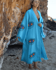 Woman wearing a turquoise Evil Eye Kaftan Robe with vibrant eye designs on a sandy beach.