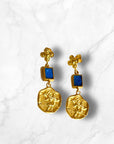 Gold earrings with blue stone accents, featuring a textured disc and elegant design on a marble background.