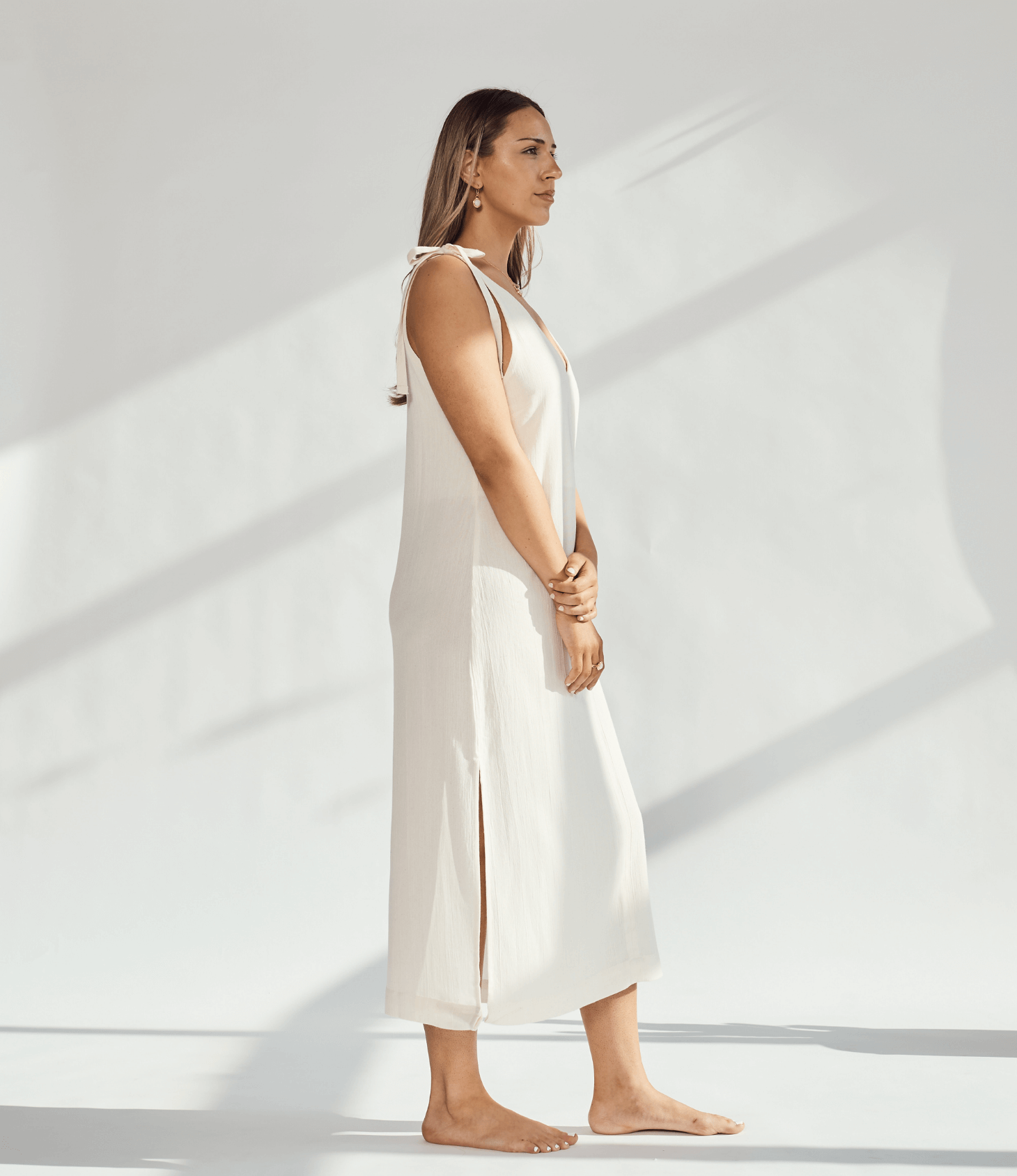 Side view of a woman in a soft cotton dress with tied straps and leg slits against a light background.