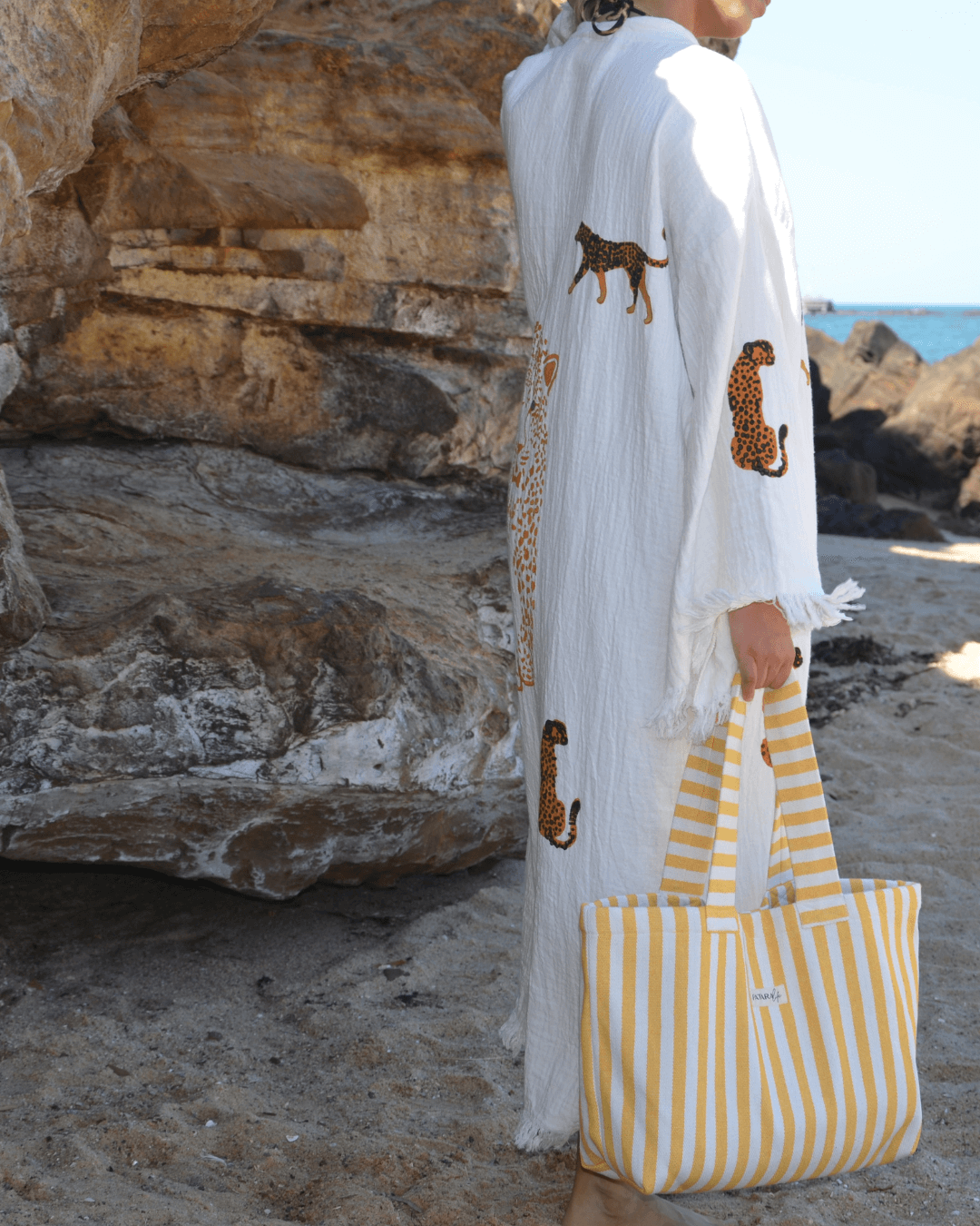 Woman wearing a Leopard Print Muslin Cotton Robe with a yellow striped tote bag at the beach.