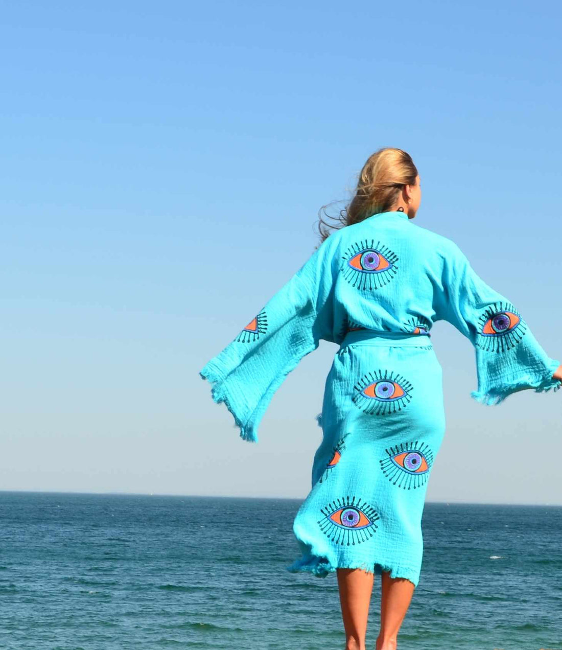 Woman wearing a turquoise Evil Eye Kaftan Robe by the ocean, showcasing unique hand-painted designs.
