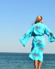 Woman wearing a turquoise Evil Eye Kaftan Robe by the ocean, showcasing unique hand-painted designs.