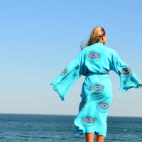 Woman wearing a turquoise Evil Eye Kaftan Robe by the ocean, showcasing unique hand-painted designs.