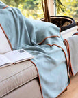 Light blue textured blanket draped over a beige sofa with an open book and a plant in the background.