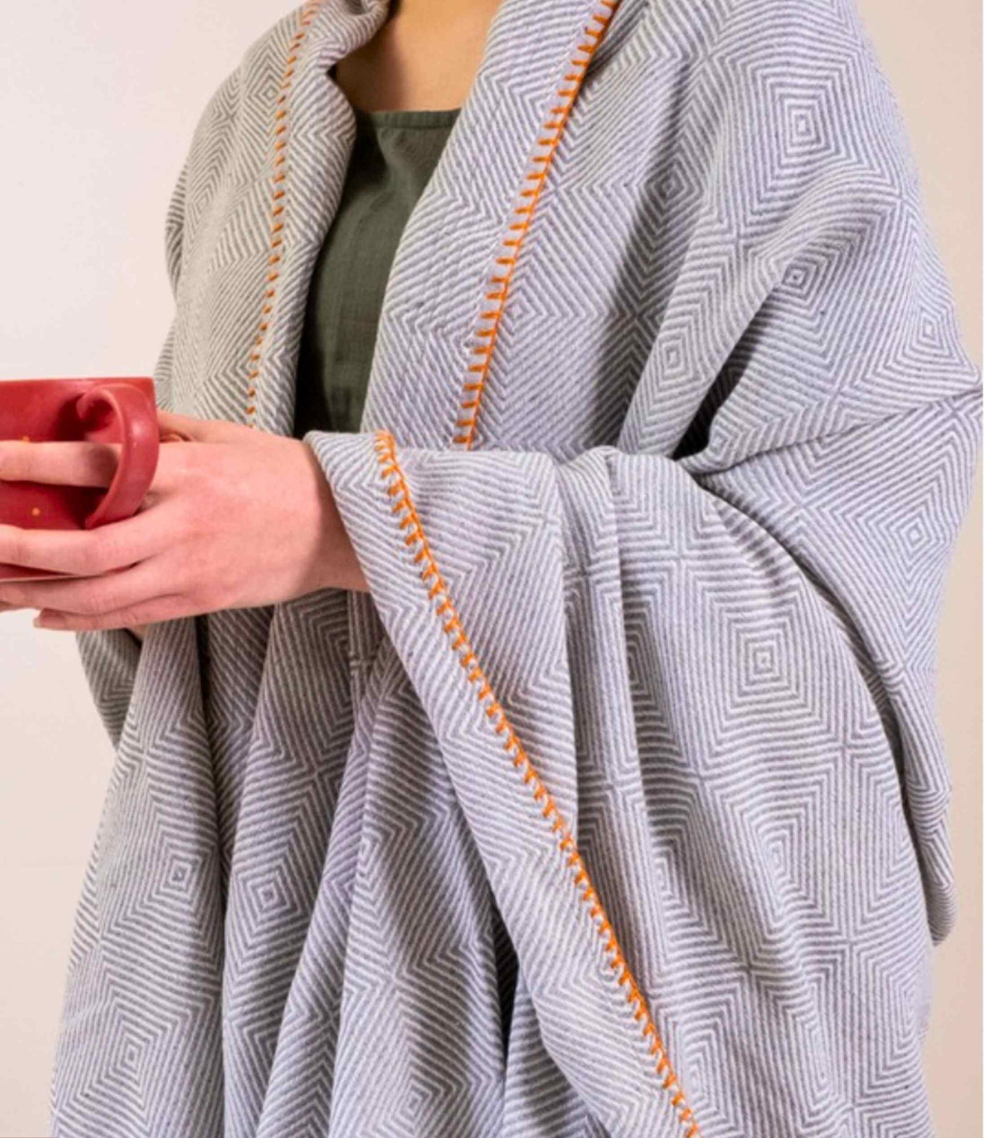Person wearing a gray patterned blanket with orange trim, holding a red mug.