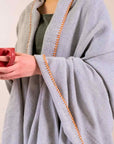 Person wearing a gray patterned blanket with orange trim, holding a red mug.