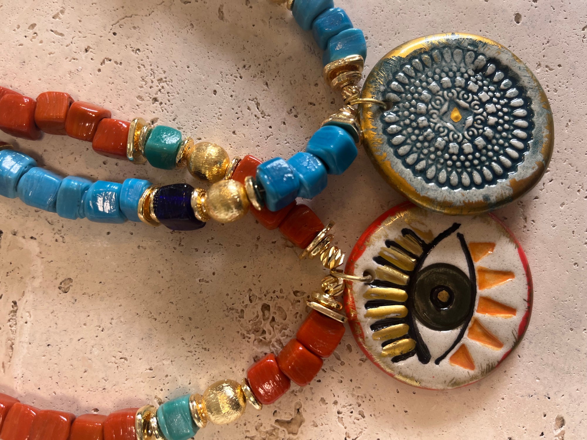 A close-up of a bohemian beaded necklace featuring terracotta red beads, turquoise blue beads, gold spacers, and two circular charms: one with a blue and gold sunburst pattern and one with an evil eye design.