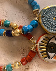 A close-up of a bohemian beaded necklace featuring terracotta red beads, turquoise blue beads, gold spacers, and two circular charms: one with a blue and gold sunburst pattern and one with an evil eye design.