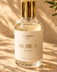 A clear glass bottle of Patara Life Aurea Room Mist with a gold cap and cream label, set on a textured beige stone surface with warm sunlight and leaf shadows.