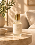 A clear glass perfume bottle with a gold cap and cream label reading 'PATARA LIFE AUREA', placed on a round stone side table in a warm, sunlit living room with a sofa and plant in the background.